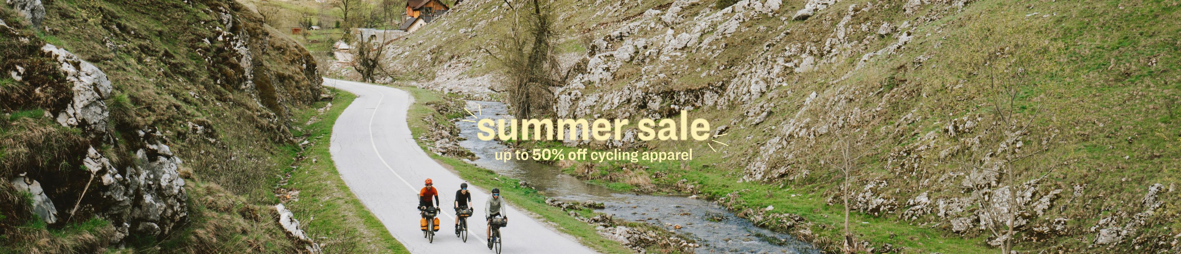 Three cyclists ride along a winding road through a scenic mountain landscape, with lush greenery and rocky hillsides. A creek runs parallel to the road, and the scene is overlaid with text reading 'summer sale up to 50% off cycling apparel.