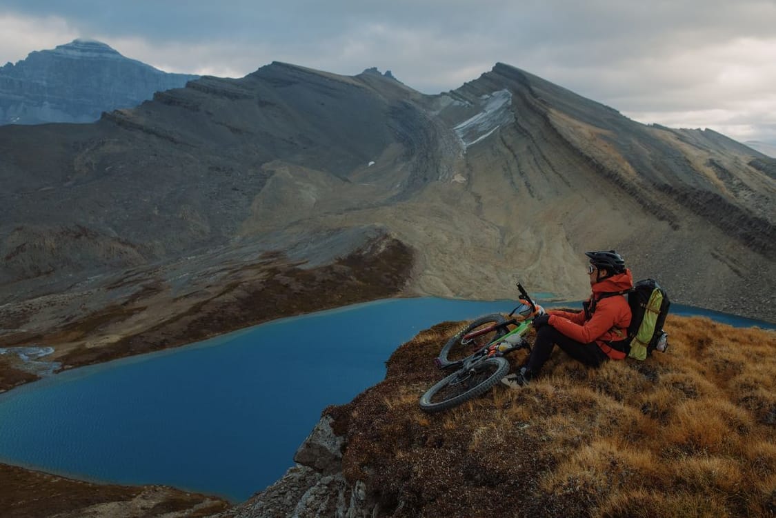 A mountain biker sits on a grassy ridge with a mountain bike beside them, overlooking a blue alpine lake and rugged rocky peaks under a cloudy sky.