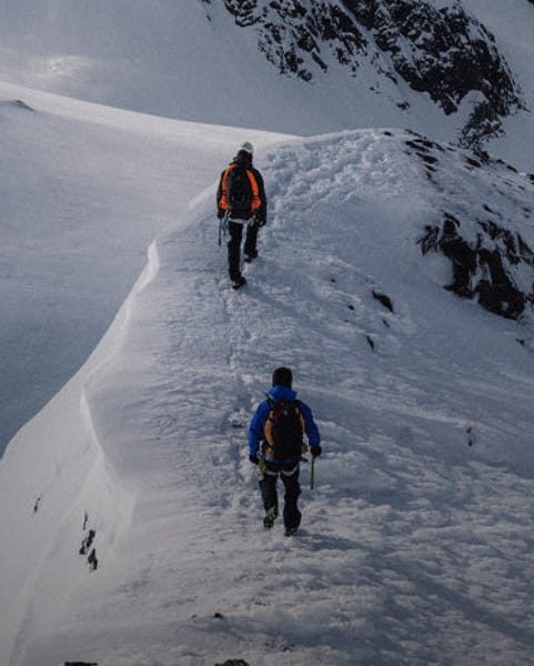 Two people hiking up a snowy mountain ridge with steep snow-covered peaks in the background.