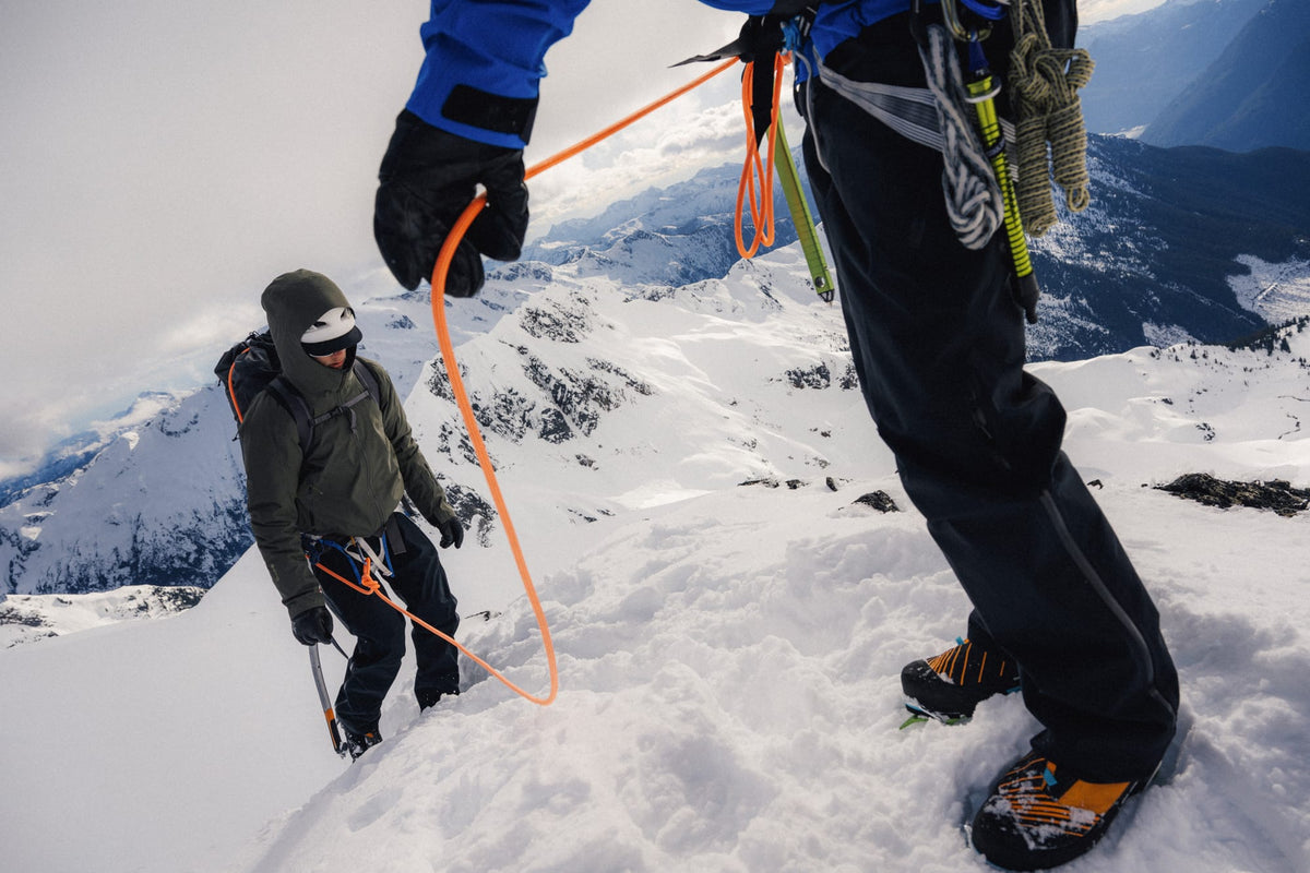 A close-up action shot of a mountaineer in a blue jacket and black pants securing an orange rope to a climber in the snow on a mountain peak, with snowy peaks in the background.