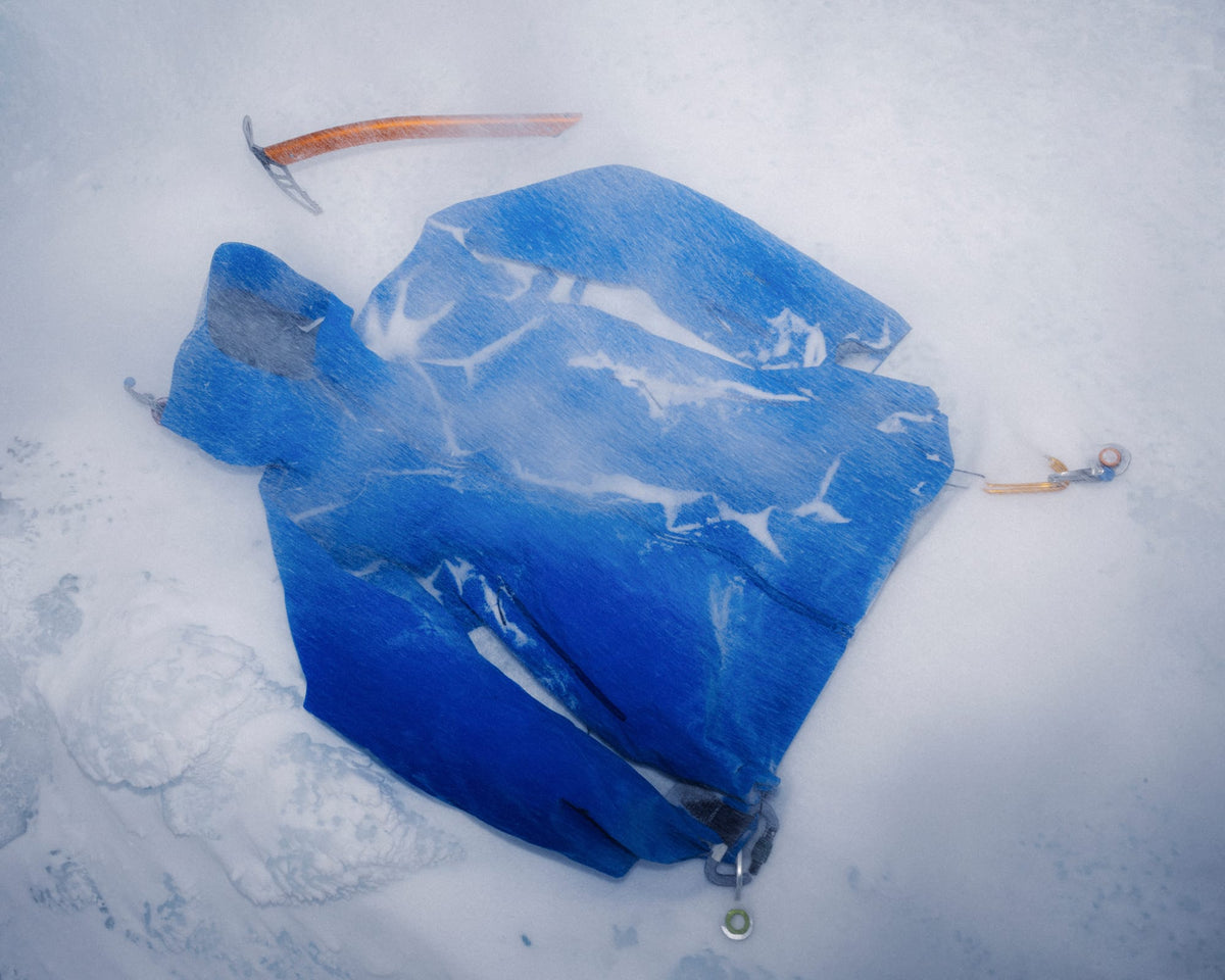 A blue ice axe lies on a snowy or icy surface, with its metal pick and orange handle visible. A second ice axe is partially visible near the top edge.