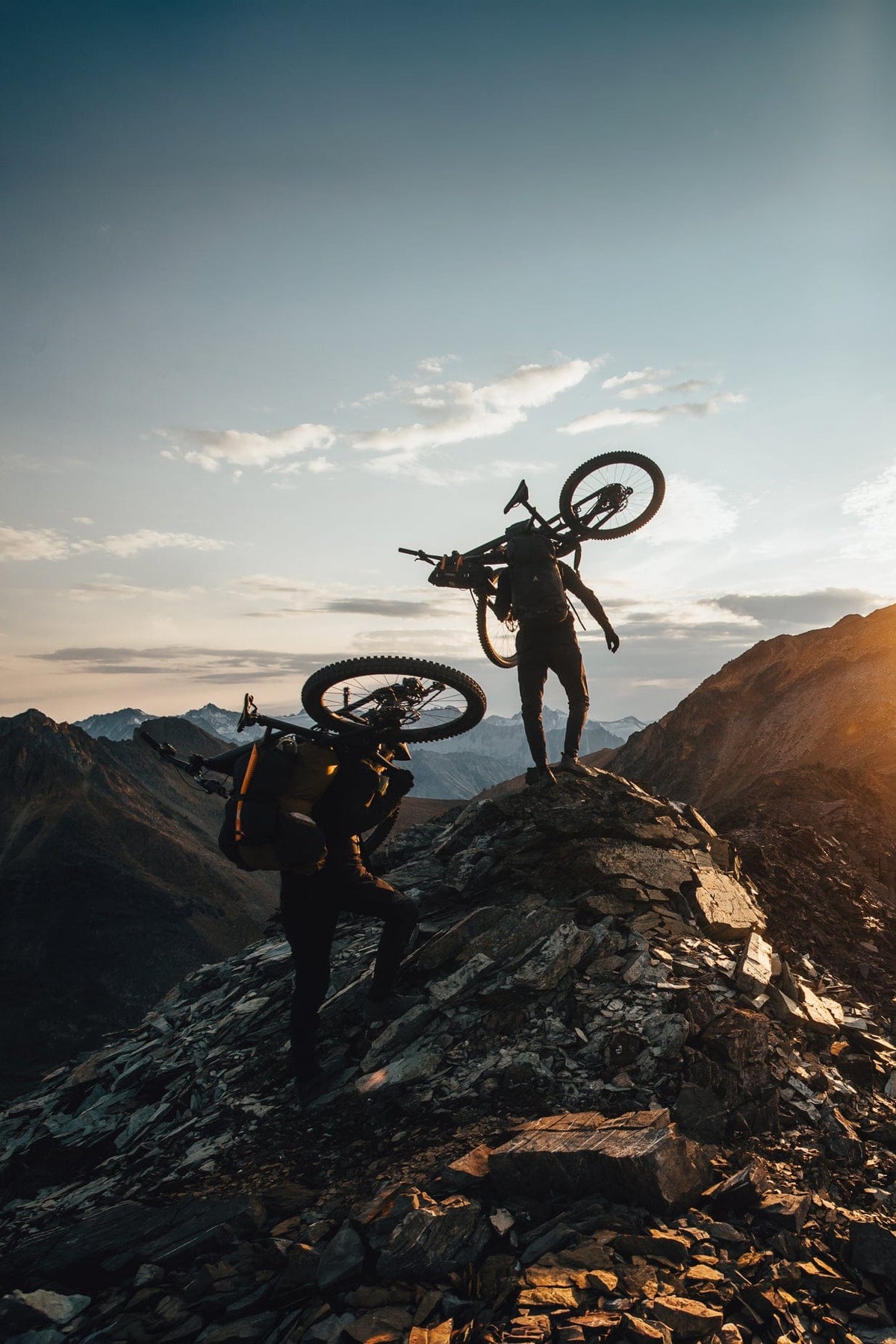 Two mountain bikers carrying bikes on rocky mountain ridges at sunset, silhouetted against a bright sun and blue sky.