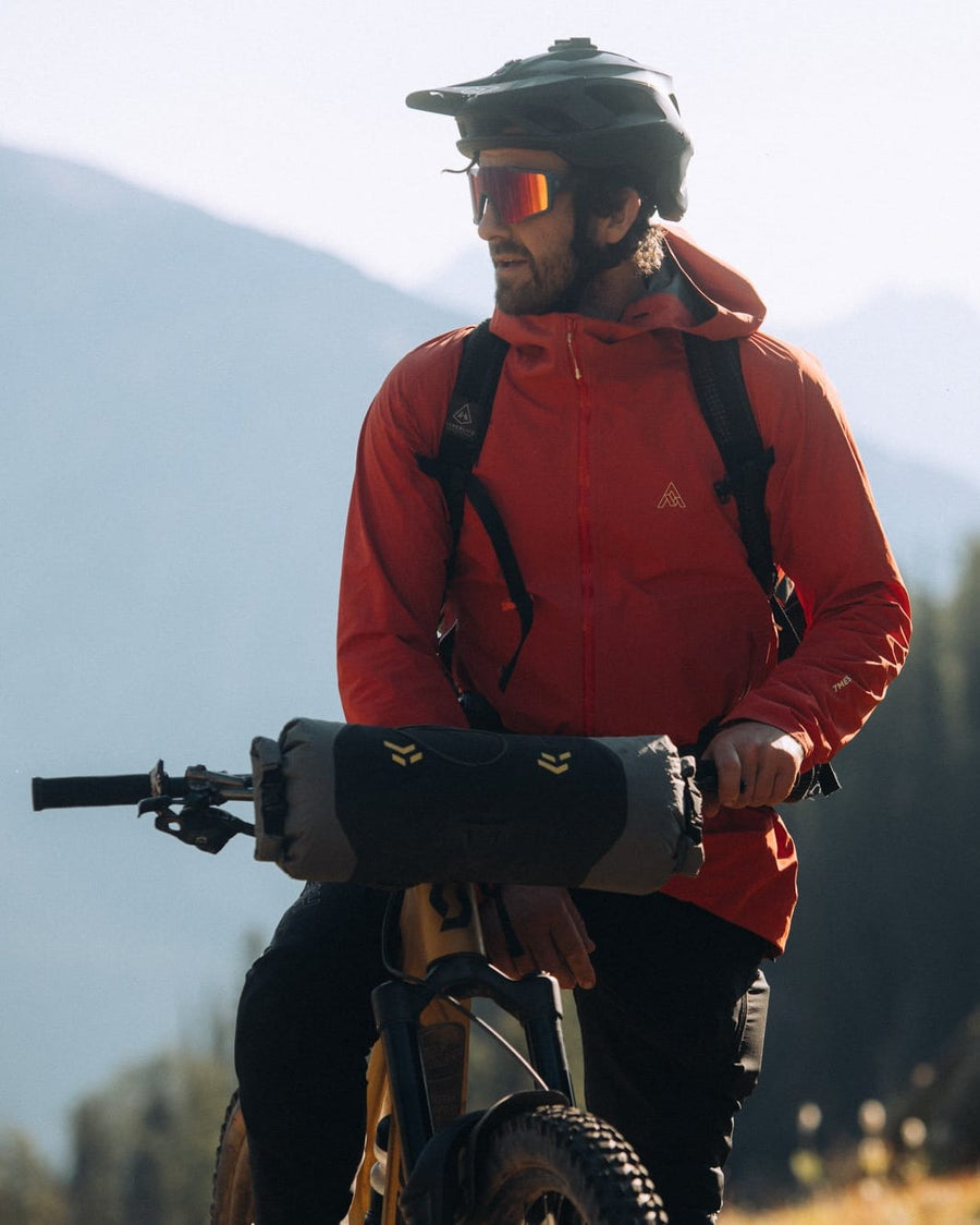 A mountain biker wearing a red hooded jacket, black helmet, and orange-tinted sunglasses stands on a trail with a mountain bike in a scenic outdoor setting with mountains and pine trees in the background.